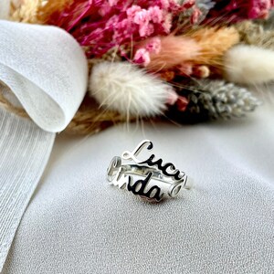 May include: A silver ring with the names "Lucy" and "Linda" in a cursive font. The ring is set against a backdrop of a bouquet of dried flowers and a white ribbon.