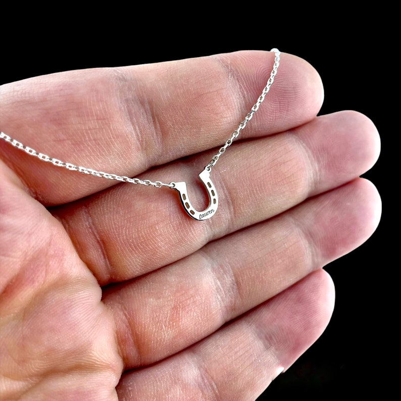 Pu&ograve; includere: Una collana d'argento a forma di ferro di cavallo con una catena delicata. Il ferro di cavallo &egrave; piccolo e presenta un design dettagliato. La collana &egrave; su una mano.