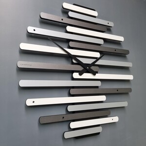 Puede incluir: Un reloj de pared moderno con un diseño único que presenta una serie de piezas rectangulares grises, blancas y negras dispuestas en un patrón escalonado. Las manecillas del reloj son negras y la esfera es minimalista.