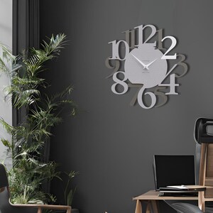 May include: A modern, silver-colored wall clock with large, stylized numerals. The clock's face is a lighter shade of silver, and the hands are visible against the background. The clock is mounted on a dark gray wall, with a plant and furniture in the room.