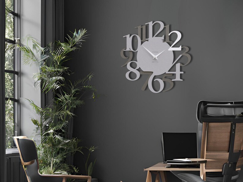 May include: A modern, silver-colored wall clock with large, stylized numerals. The clock's face is a lighter shade of silver, and the hands are visible against the background. The clock is mounted on a dark gray wall, with a plant and furniture in the room.