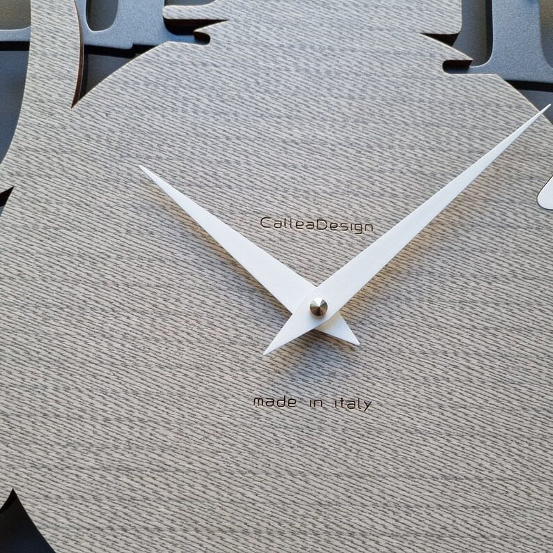 May include: A modern wall clock with a light gray wood grain texture. The clock features white hands and the text "CalleaDesign" and "made in italy". The clock's shape is a stylized tree.