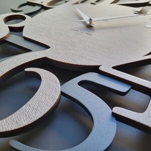 May include: A modern wall clock with a light wood grain face and gray numerals. The clock hands are white, and the text "made in italy" is visible. The numbers are cut out, creating a layered effect. The clock is a decorative home decor item.