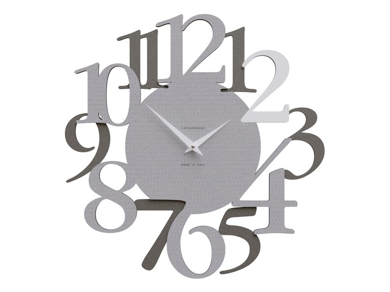 May include: A modern wall clock with a gray textured face and cut-out numbers in varying shades of gray. The clock hands are white, and the text "CarlaDesign" and "made in italy" are visible.