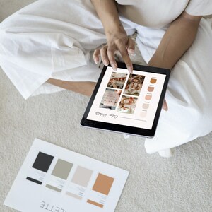 May include: A person is reviewing a color palette on a tablet. The tablet screen shows a color palette with different shades of brown and beige. The person is sitting on a carpet and has a piece of paper with a color palette on it.