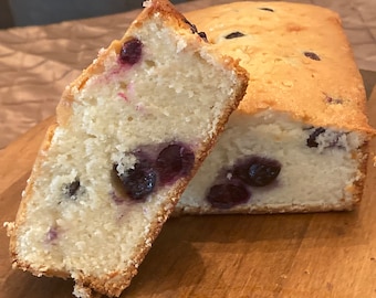 May include: A loaf of blueberry bread sliced in half, revealing the blueberries inside. The bread is light brown and has a slightly golden crust. The bread is sitting on a wooden cutting board.