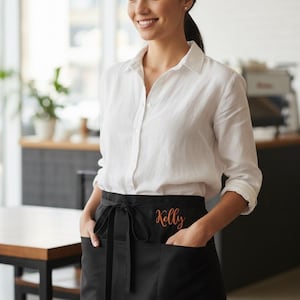 May include: A black waist apron with two front pockets and a tie-waist. The apron has the name "Kelly" embroidered in orange script. The person is wearing a white button-down shirt.
