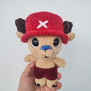 May include: A plush, handmade character doll with a red hat featuring a white cross, brown antlers, and dark red shorts. The doll has large black eyes, a blue nose, and tan fur. The doll is held in a person's hand.