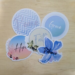 Peut inclure: Cinq autocollants ronds avec des motifs et des mots différents. Les autocollants sont bleus, bleu clair, roses, blancs et une fleur bleue aquarelle. Les mots sur les autocollants sont "Paix", "Grâce", "Espoir", "Foi" et "Patience".