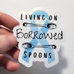 May include: A blue ribbon with two spoons crossed over it. The text "Living on Borrowed Spoons" is written on the ribbon.