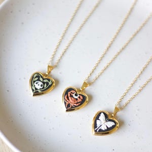 Puede incluir: Tres collares de oro con colgantes en forma de corazón con diferentes diseños. El primer colgante presenta una calavera y enredaderas, el segundo un cangrejo y una luna creciente, y el tercero una mariposa.