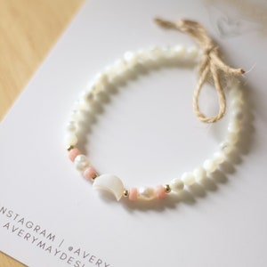 May include: A white and pink beaded bracelet with a gold crescent moon charm. The bracelet is tied with a brown string.