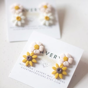 Puede incluir: Un par de pendientes colgantes con dos flores de margarita blancas y amarillas con un centro marrón, y dos flores de girasol amarillas y marrones con un centro marrón. Los pendientes se muestran en una tarjeta blanca con el texto "Avery May" y "Instagram | @avery.may.designs averymaydesigns.etsy.com".