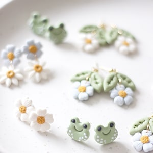 Puede incluir: Un conjunto de pendientes de arcilla polimérica con varios diseños, incluyendo margaritas, hojas y ranas. Los pendientes son de color verde claro, blanco y amarillo.