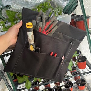 Puede incluir: Un organizador negro de herramientas de jardinería con múltiples bolsillos, que contiene guantes de jardinería, una paleta, unas tijeras de podar y un bolígrafo. El organizador está diseñado para guardar herramientas de jardinería. El fondo muestra un invernadero con plantas en macetas rojas.