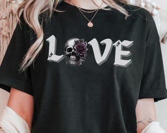 Könnte beinhalten: Schwarzes T-Shirt mit dem Wort "LOVE" in weißen, 3D-Buchstaben. Das "O" wird durch einen Totenkopf mit einer lila Rose ersetzt. Das Shirt hat kurze Ärmel und einen Rundhalsausschnitt.