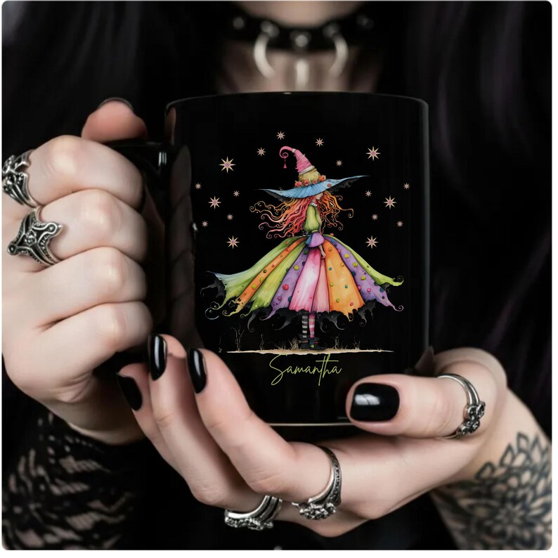 Puede incluir: Taza de cer&aacute;mica negra con una colorida ilustraci&oacute;n de acuarela de una bruja con un vestido y sombrero caprichosos. El nombre de la bruja, Samantha, est&aacute; escrito debajo. La taza es sostenida por una persona con esmalte de u&ntilde;as negro.