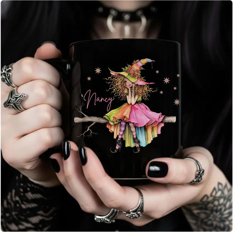 Puede incluir: Taza de cer&aacute;mica negra con una colorida ilustraci&oacute;n de una bruja sentada en una rama. La bruja lleva un vestido y un sombrero arco&iacute;ris, con el nombre "Nancy" en la taza. Estrellas dispersas alrededor de la bruja.