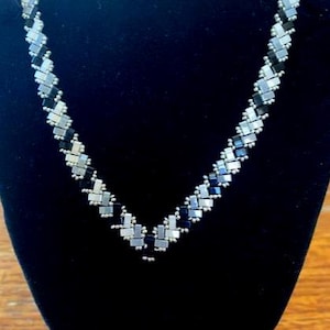 Può includere: Una collana geometrica con un design a forma di V. La collana presenta un motivo di perline rettangolari argentate e nere alternate. La collana è esposta su un busto di velluto nero.