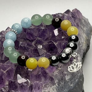 Op de afbeelding: Een kralenarmband met diverse gekleurde stenen, waaronder blauw, groen, geel en zwart, wordt getoond op een paarse amethistgeode. Een zilveren lotusbloem bedel hangt aan de armband. Dit is een sieraad.