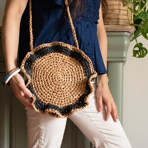 May include: A round crocheted bag with a brown and black striped border. The bag has a scalloped edge and is being held by a person.