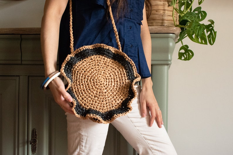 May include: A round crocheted bag with a brown and black striped border. The bag has a scalloped edge and is being held by a person.