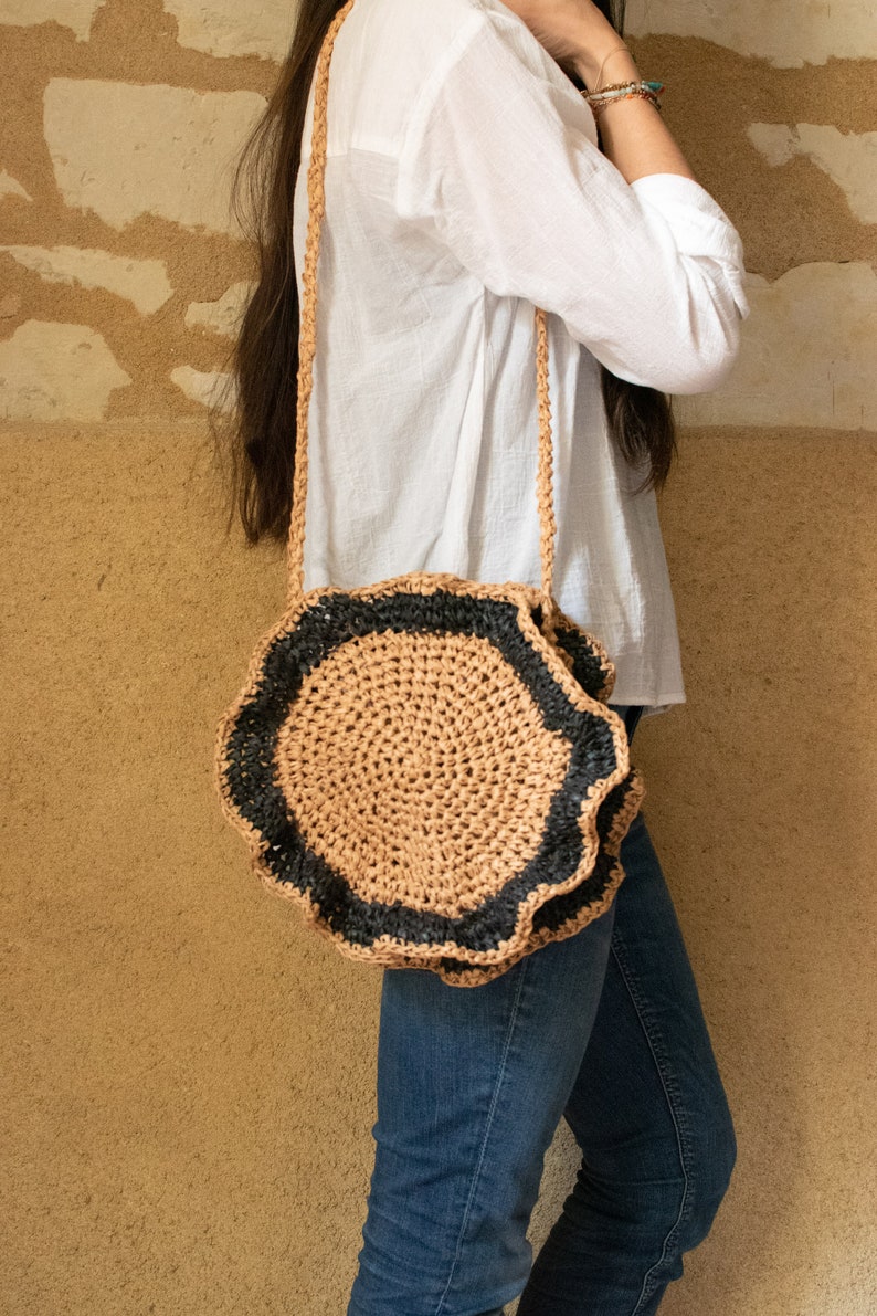 May include: A round crocheted bag with a black and brown design. The bag has a long strap and is worn over the shoulder.