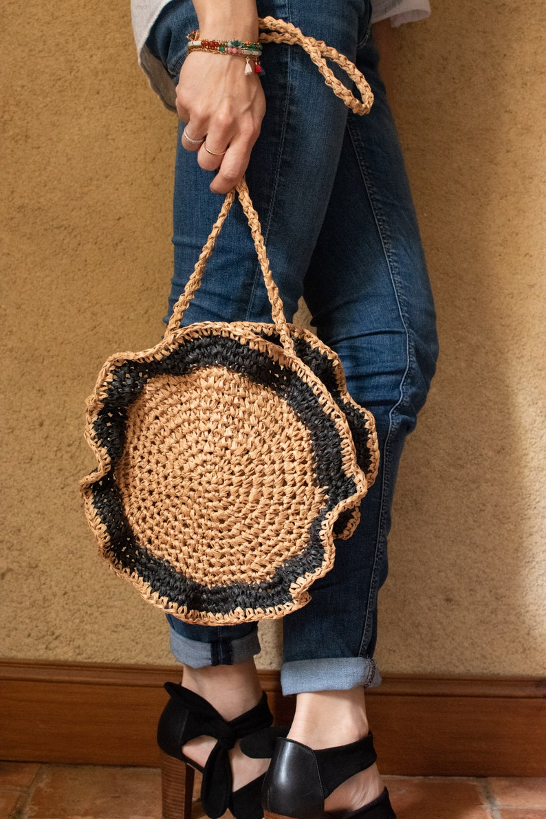 May include: A round straw bag with a black crocheted trim. The bag has a long strap and is being held by a person wearing blue jeans.