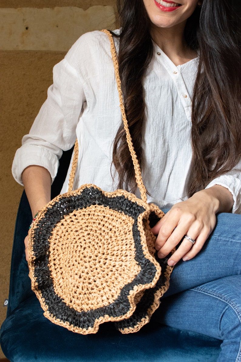 May include: A round, crocheted bag with a black trim and a ruffled edge. The bag is made of natural fibers and has a long strap.