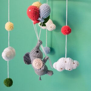 May include: A gray crocheted elephant with pink ears and a white crocheted cloud with gold stars hanging from a mobile. The mobile also features a red, yellow, and blue crocheted balloon.