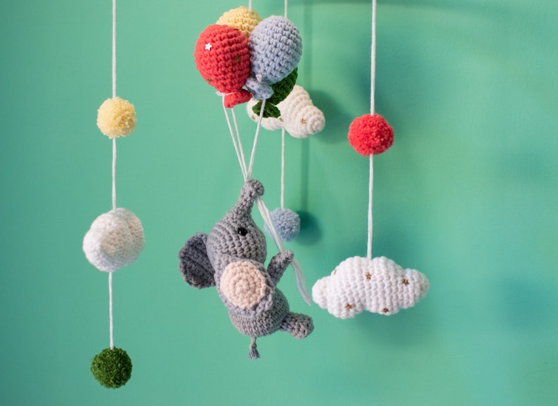 May include: A gray crocheted elephant with pink ears and a white crocheted cloud with gold stars hanging from a mobile. The mobile also features a red, yellow, and blue crocheted balloon.
