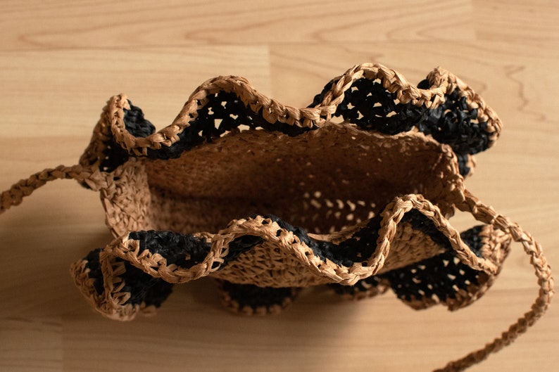 May include: A brown and black woven bag with a ruffled edge. The bag has a single strap and is made of a natural material.