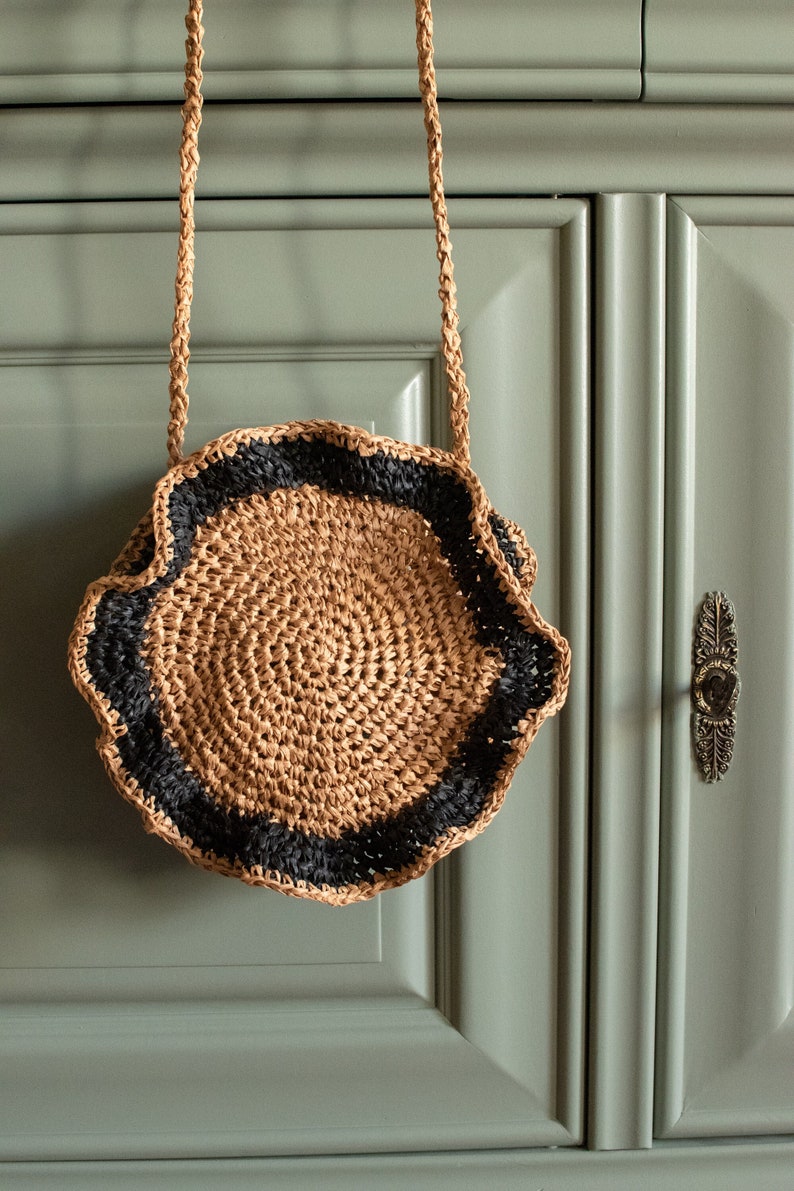 May include: A round, crocheted straw bag with a black trim. The bag has a long strap and is hanging on a green cabinet.