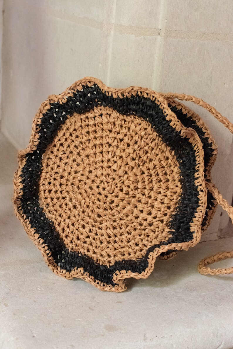 May include: A round straw bag with a black trim and a scalloped edge. The bag has a woven texture and a long strap.