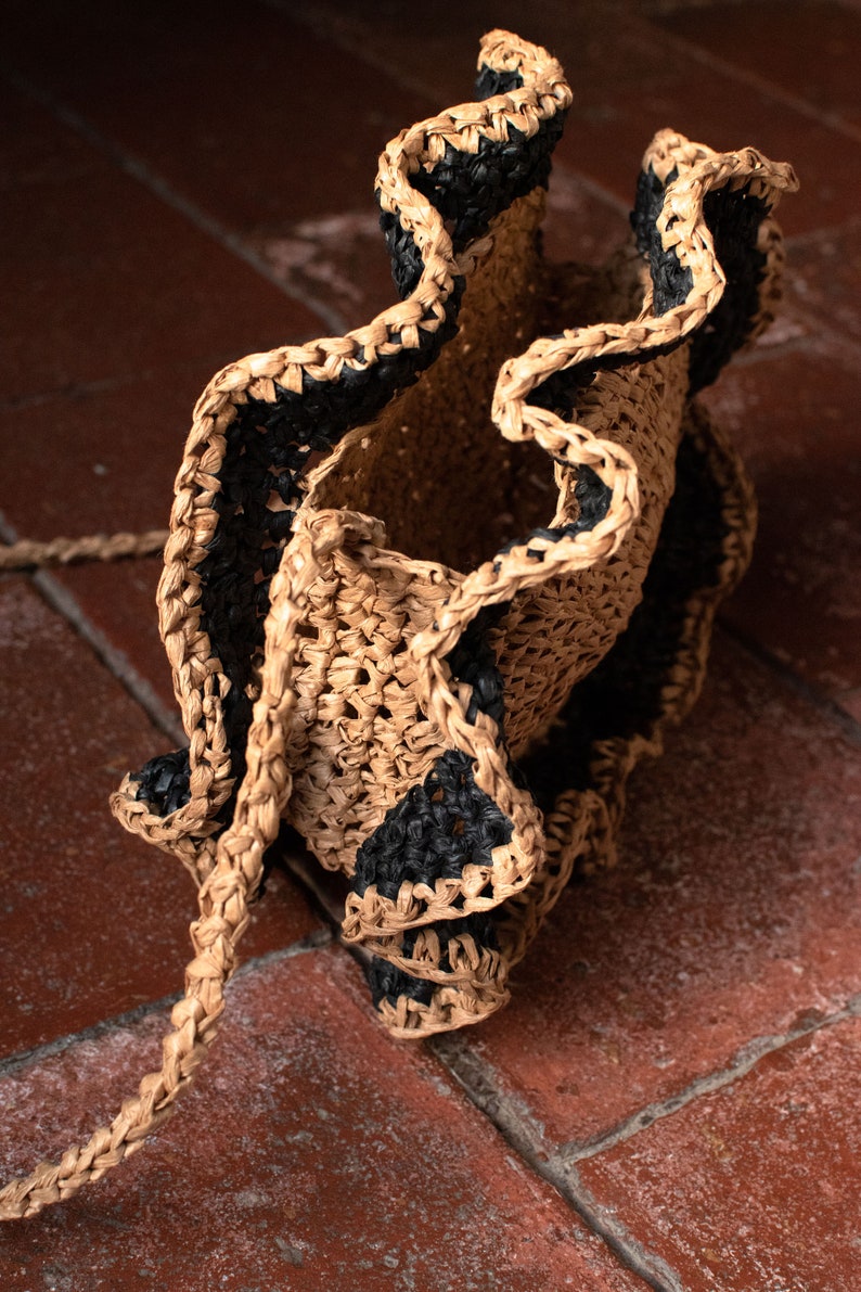 May include: A woven straw bag with black trim. The bag has a ruffled design and is made of natural materials.