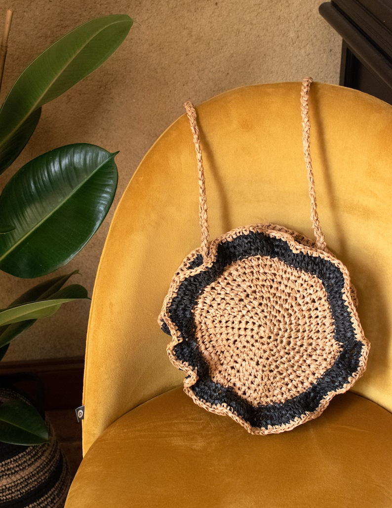 May include: A round, crocheted bag with a black trim and a scalloped edge. The bag has two long straps and is sitting on a yellow chair.