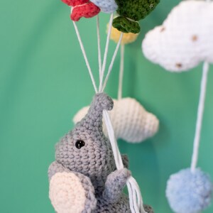 May include: A gray crocheted elephant toy hanging from a mobile with colorful crocheted balloons and clouds. The elephant has a pink ear and a black eye.