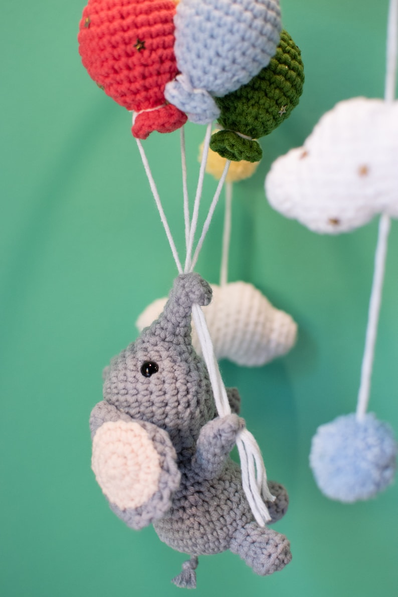 May include: A gray crocheted elephant toy hanging from a mobile with colorful crocheted balloons and clouds. The elephant has a pink ear and a black eye.