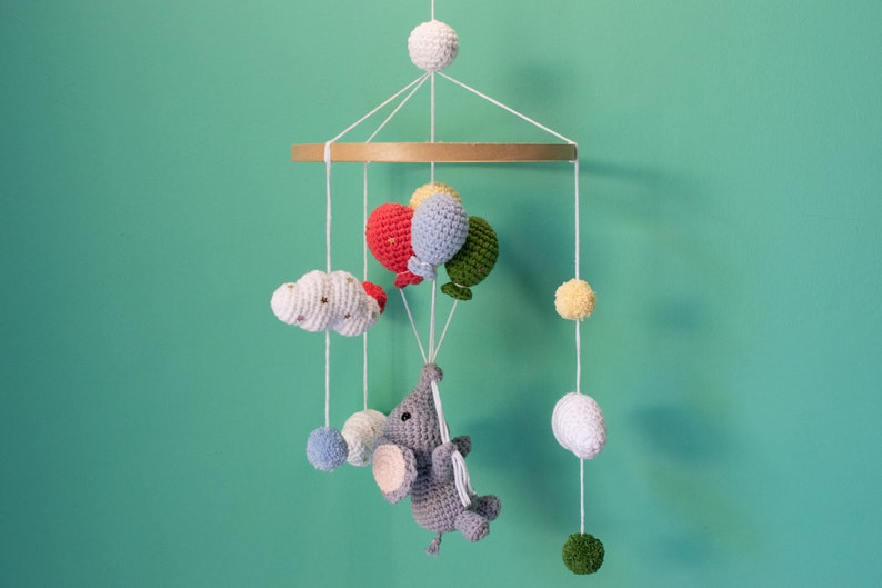 May include: A crocheted baby mobile with a gray elephant holding balloons. The balloons are red, blue, green, and yellow. The mobile also has white clouds and pom-poms in various colors.