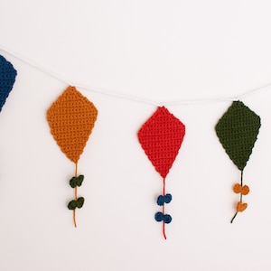 Puede incluir: Una guirnalda de cometas de crochet en azul, naranja, rojo, verde y gris, con pequeñas flores de crochet colgando debajo de cada cometa.