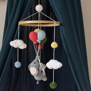 May include: A crocheted baby mobile with a gray elephant holding balloons, white clouds, and colorful balls hanging from a wooden ring.