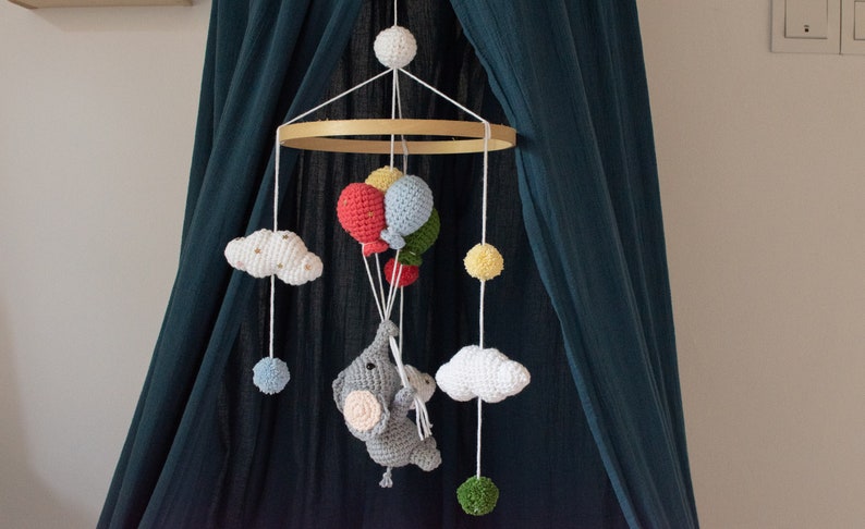 May include: A crocheted baby mobile with a gray elephant holding balloons, white clouds, and colorful balls hanging from a wooden ring.