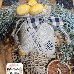 May include: A small burlap sack filled with yellow, speckled faux potatoes. The sack is tied with a black and white checkered ribbon and features a rectangular tag with the word "Potatoes" written in cursive script and a small checkered patch.