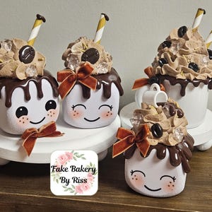 May include: A collection of whimsical, hand-painted ceramic coffee cup decorations. Each cup features a smiling face, chocolate drizzle, whipped cream topping, coffee beans, and a decorative straw. A small bow adds a touch of elegance.
