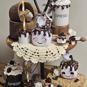 May include: A tiered wooden tray decorated with a coffee and marshmallow theme. The tray features a variety of ceramic mugs, including a white mug with the word "ESPRESSO" and a black mug with the word "ESPRESSO". The tray also includes a variety of decorative elements, such as chocolate-covered marshmallows, coffee beans, and graham crackers.