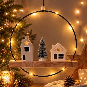 May include: A circular black metal hoop wrapped with warm white fairy lights, featuring a horizontal wooden shelf suspended inside. On the shelf sit two small white ceramic houses and a miniature green pine tree, all illuminated by the lights.
