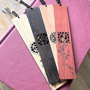 May include: Four wooden bookmarks with intricate designs and tassels rest on a pink book. The bookmarks vary in color from light beige to dark brown and red, each featuring unique cut-out patterns and etched details. The tassels are black with wooden beads.