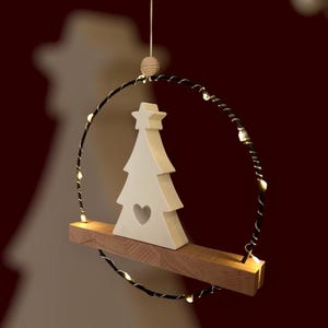 Könnte beinhalten: Ein weißer Weihnachtsbaum aus Keramik mit einem Herz-Ausschnitt, der auf einem Holzregal steht. Das Regal ist in einem schwarzen Drahtring aufgehängt, der mit kleinen, warmweißen Lichtern verziert ist. Oben befinden sich eine Holzperle und eine Schnur.