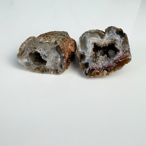 May include: Two natural geode halves, showcasing a hollow interior filled with crystal formations. The outer shell displays a mix of brown, gray, and white hues. The geodes are set against a white background, highlighting their unique textures and colors.
