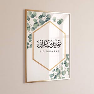 May include: A gold framed print with a white background and a gold hexagon in the center. The hexagon contains Arabic calligraphy that reads "Eid Mubarak" in English. The hexagon is surrounded by green watercolor eucalyptus leaves.